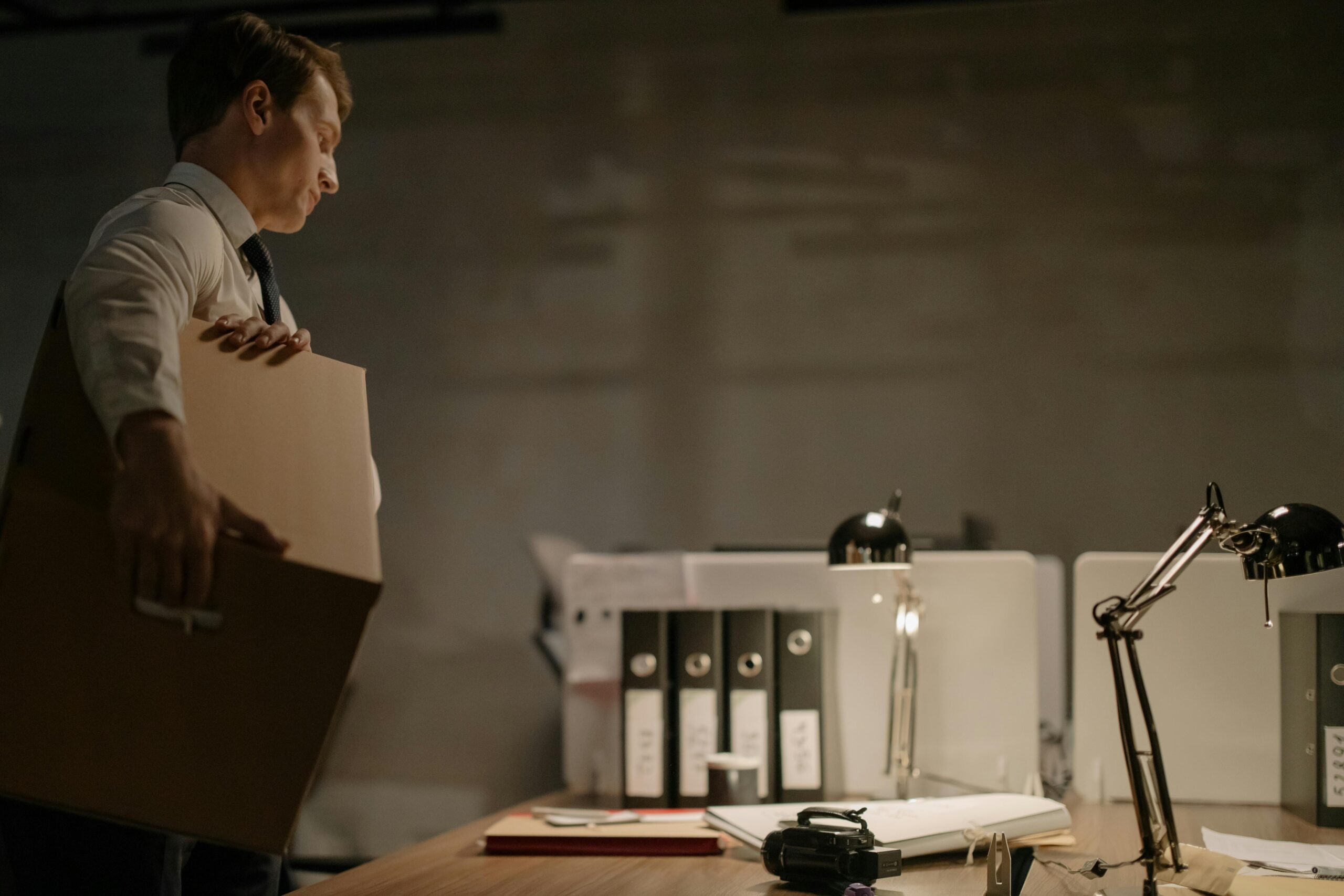 Businessman stands at desk packing documents in a dimly lit office setting.